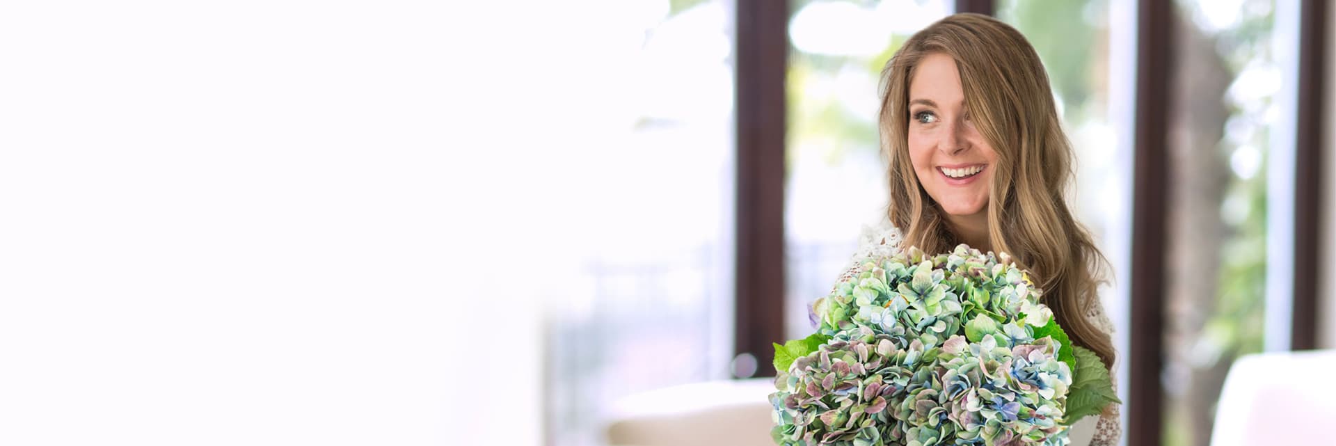A joyful woman holding a large bouquet of blue and green hydrangeas indoors.